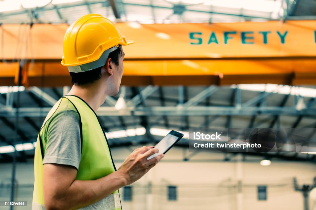Young Asian male factory worker using a digital tablet Young Asian male heavy industral worker using a digital tablet inside manufacturing and factory site with crane behind Safety Stock Photo