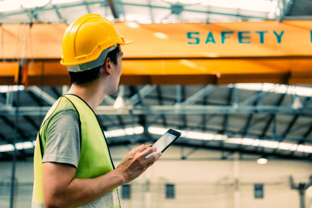 Young Asian male factory worker using a digital tablet Young Asian male heavy industral worker using a digital tablet inside manufacturing and factory site with crane behind safety stock pictures, royalty-free photos & images