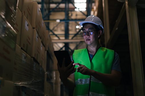 Young Asian male warehouse worker using a digital tablet inside warehouse to check inventory and stock management in factory - storage and factory stocktake management concept