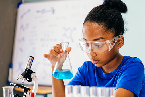 Young African American mixed kid testing chemistry lab experiment and shaking glass tube flask along with microscope and whiteboard in background - science and learning education concept