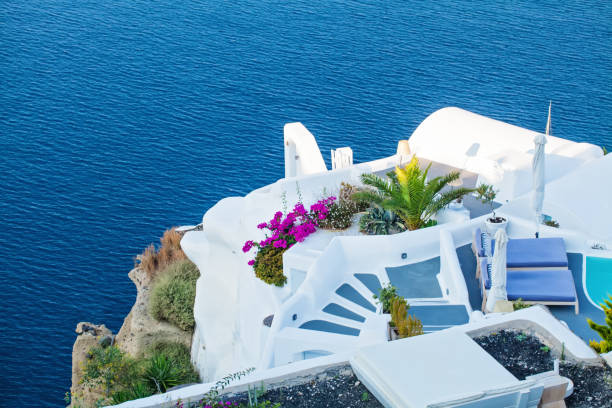 Bask in the beauty of Santorini's idyllic landscapes with this stunning view from a sunlit terrace. The white architecture harmonizes with the azure sea, creating a picturesque scene of Mediterranean splendor. Adorned with vibrant flowers and potted cacti, the terrace offers a tranquil retreat amidst the island's breathtaking beauty. From this vantage point, guests can soak in the panoramic views of the coastal landscape, immersing themselves in the essence of Santorini's charm and serenity on a sunny summer day.