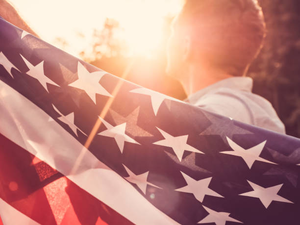 Handsome, young man waving an American flag Handsome man waving an American flag against a background of trees and blue sky. View from the back, close-up. National holiday concept patriotic man stock pictures, royalty-free photos & images