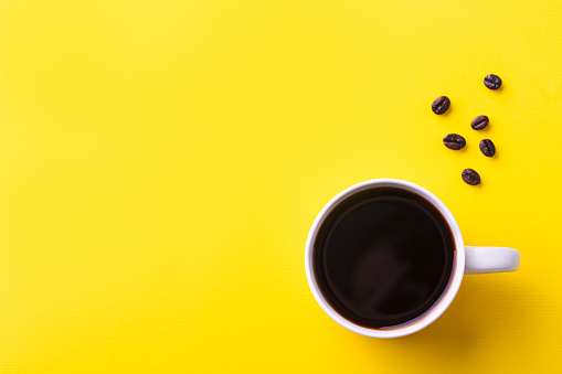 Cup of black coffee with coffee beans on yellow background. Top view. Copy space
