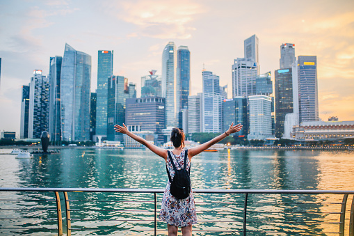 Young traveler woman with her arms outstretched is watching sunset above the amazing skyline in Singapore bay area. Freedom, travel, concepts.