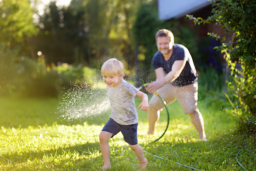 Funny little boy with his father playing with garden hose in sunny backyard. Preschooler child having fun with spray of water. Summer outdoors activity for kids.