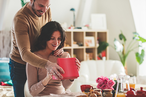 Smiling mid adult man surprising his girlfriend with a gift she is opening on a romantic date.