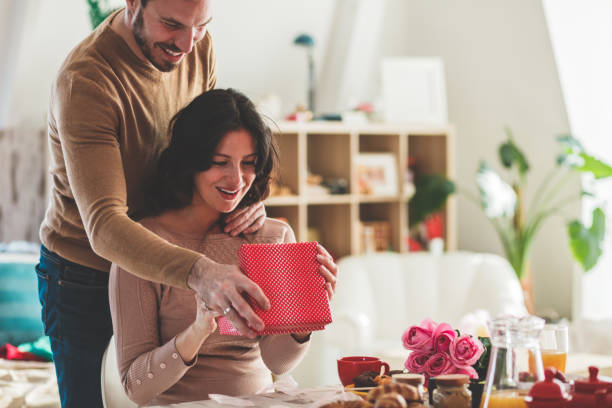 Giving her a gift Smiling mid adult man surprising his girlfriend with a gift she is opening on a romantic date. valentines present stock pictures, royalty-free photos & images