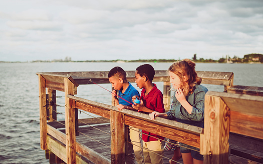 Group of boys and girls on a dock or pier with a fishing rod play and have fun trying to catch a fish together. Elementary age friends in casual clothes outdoors in the summer at the ocean or lake or big body of water trying their best to land a catch