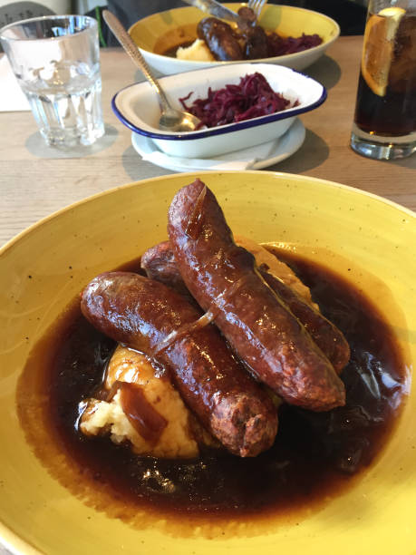 Stock photo of homemade bangers and mash being eaten at wooden dining table for pub lunch with fried onions and rich gravy, sausages and mashed potato recipe, eating processed meat. imagen de pandilleros británicos tradicionales y receta de puré/salchicha y puré, puré de patatas, salsa de cebolla, restaurante gourmet pub almuerzo, comida casera abundante, carne de res, venado y salchichas de cerdo/bratwursts alemanas, cocina frita