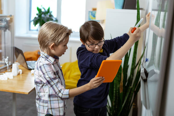 istock Blonde boy carrying orange tablet while his friend writing on the desk 1148332655