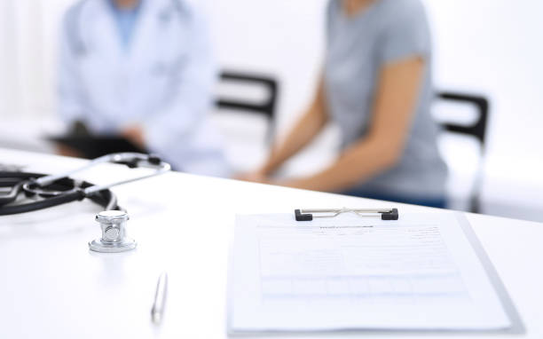 istock Stethoscope, clipboard with medical form lying on hospital reception desk with laptop computer and busy doctor and patient communicating at the background. Medical tools at doctor working table.Medicine concept 1148338238