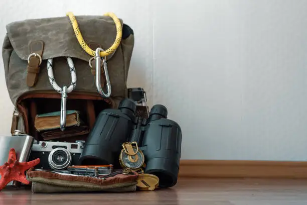 Travel or adventure background. A backpack with a safety rope and carabines, binoculars, compass, book and photo camera on a floor on a white wall background.