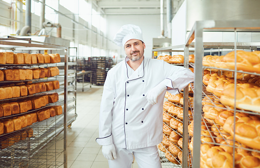 A baker, a man in white uniform, is standing in a bakery against the shelves of bread.