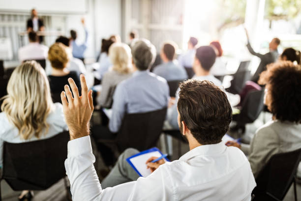 Back view of a businessman raising his hand on a seminar. Rear view of a businessman raising his hand to ask the question on a seminar with large group of his colleagues. convention center stock pictures, royalty-free photos & images