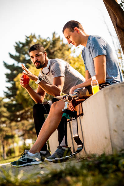 istock Two active, multi-ethnic buddies, one of them with an artificial sports leg prosthetic, talking and hydrating themselves with energy drinks while sitting on a park bench 1148381712