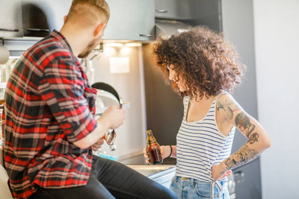 Showing his flatmate opening bottle with spoon Young woman having bottled beer and laughing with her male flatmate sitting on kitchen counter flatmate stock pictures, royalty-free photos & images