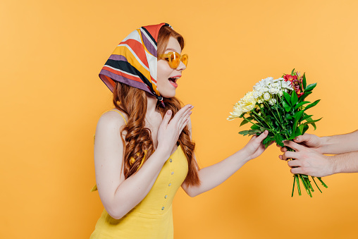 surprised stylish girl in headscarf and sunglasses gesturing and looking at flowers isolated on yellow