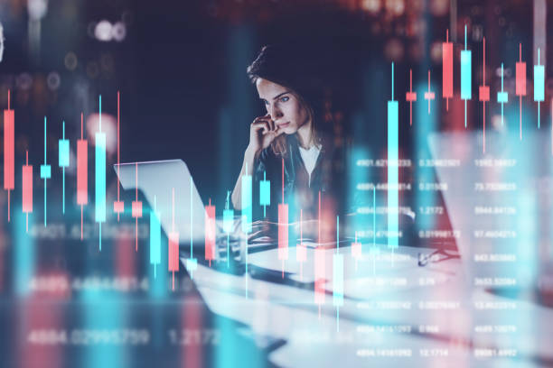 Business woman sitting at night office in front laptop computer with financial graphs and statistics on monitor. Red and green candlestick chart and stock trading on background. Double exposure. stock photo