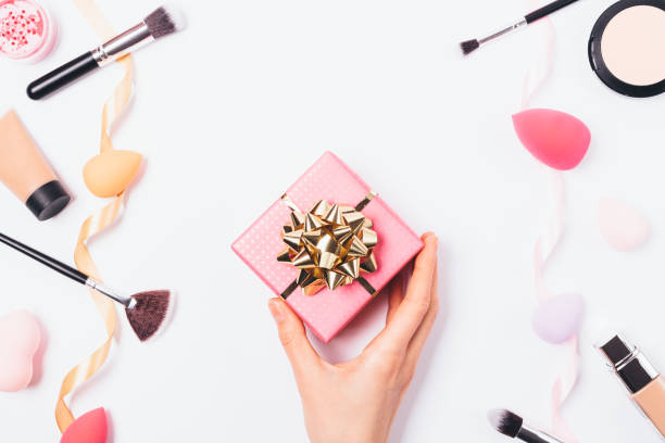 Woman's hand holding pink gift box Woman's hand holding pink gift box among decorative cosmetics and accessories on white table, top view. Flat lay composition of makeup products, professional brushes and sponges. person holding frame over face stock pictures, royalty-free photos & images