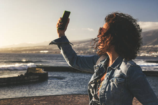 schöne mädchen, die selfie, warmes licht des sonnenuntergangs abend woman video-anruf mit smartphone im freien, ozeanwellen im hintergrund female mit windigen haaren mit technologie, um überall verbunden zu bleiben - disply stock-fotos und bilder