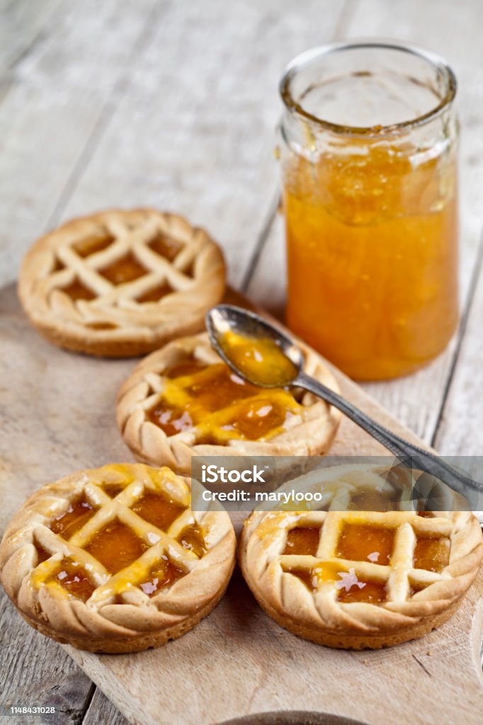 Fresh baked tarts with marmalade filling and apricot jam in glass jar on cutting board on rustic wooden table. Fresh baked tarts with marmalade filling and apricot jam in glass jar on cutting board on rustic wooden table background. Preserves Stock Photo