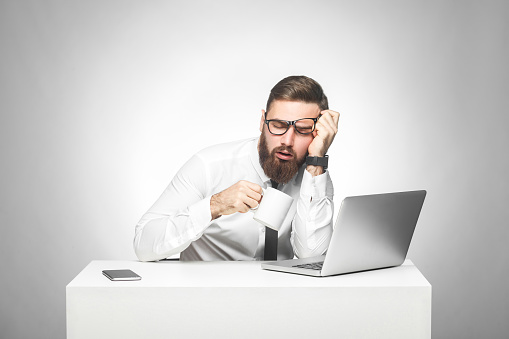 Irresponsible tired young manager in white shirt and black tie are sitting in office and trying not to sleep on the work, drink a cup of coffee, holding head with hand. Studio shot, isolated, indoor