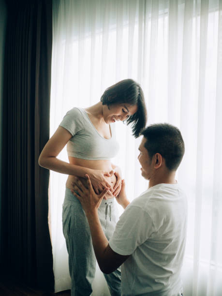 アジアの妊娠カップルが自宅のリビングルームで腹を触って聞いています。 - rubbing human hand togetherness women ストックフォトと画像
