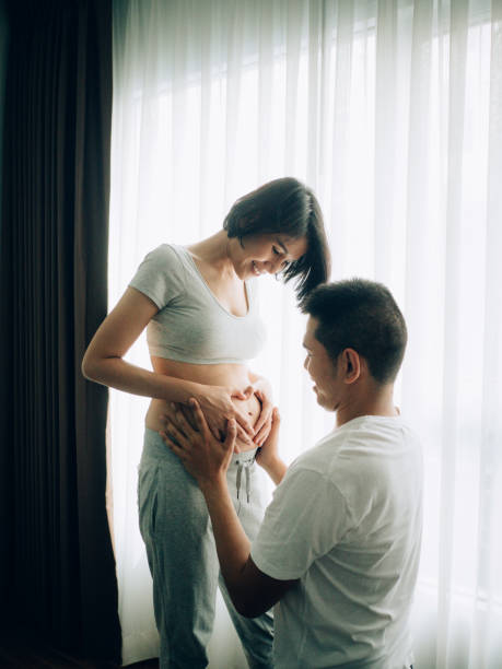 couple enceinte asiatique touchant et écoutant le ventre dans le salon à la maison. - rubbing human hand togetherness women photos et images de collection