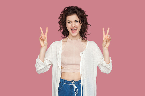 Portrait of happy funny excited beautiful brunette young woman with curly hairstyle in casual style standing with victory or v sigg, looking at camera. indoor studio shot isolated on pink background.