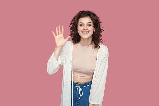 Hi, glad to see you. Portrait of happy satisfied beautiful brunette young woman with curly hairstyle in casual style standing, looking at camera and greeting. studio shot isolated on pink background.