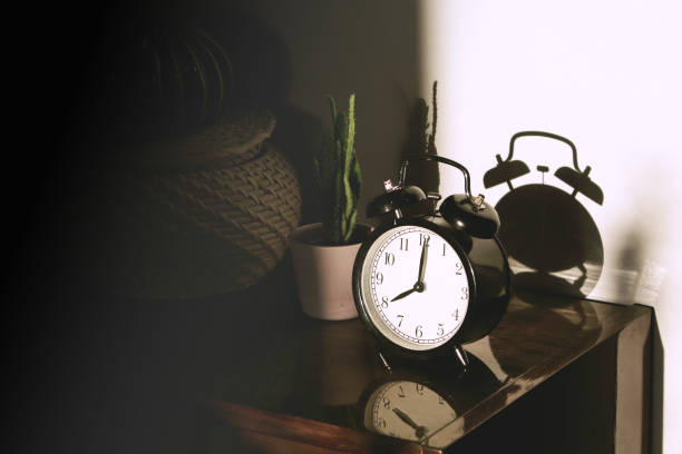 istock Black retro vintage alarm clock on a wooden bedside table with reflection in a polished surface in the rays of the setting sun with hard shadows 1148450880