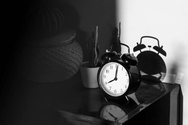 istock Black retro vintage alarm clock on a wooden bedside table with reflection in a polished surface in the rays of the setting sun with hard shadows 1148450906