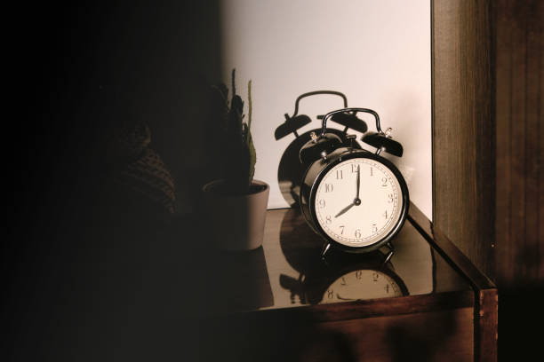 istock Black retro vintage alarm clock on a wooden bedside table with reflection in a polished surface in the rays of the setting sun with hard shadows 1148451408
