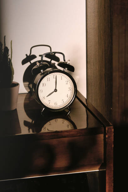 istock Black retro vintage alarm clock on a wooden bedside table with reflection in a polished surface in the rays of the setting sun with hard shadows 1148451425