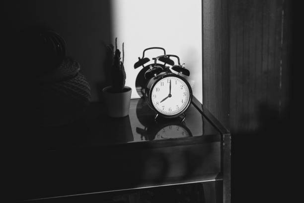 istock Black retro vintage alarm clock on a wooden bedside table with reflection in a polished surface in the rays of the setting sun with hard shadows 1148451436