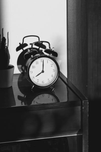 istock Black retro vintage alarm clock on a wooden bedside table with reflection in a polished surface in the rays of the setting sun with hard shadows 1148451452