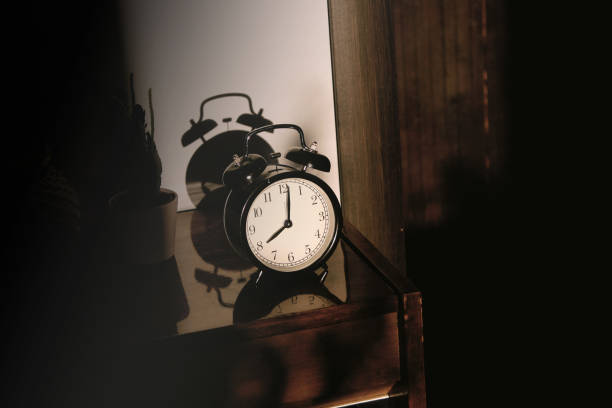 istock Black retro vintage alarm clock on a wooden bedside table with reflection in a polished surface in the rays of the setting sun with hard shadows 1148451459