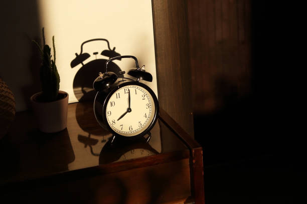 istock Black retro vintage alarm clock on a wooden bedside table with reflection in a polished surface in the rays of the setting sun with hard shadows 1148451795