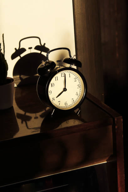 istock Black retro vintage alarm clock on a wooden bedside table with reflection in a polished surface in the rays of the setting sun with hard shadows 1148451976