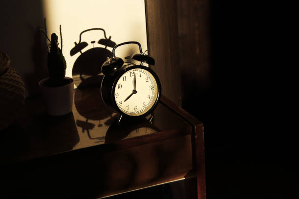 istock Black retro vintage alarm clock on a wooden bedside table with reflection in a polished surface in the rays of the setting sun with hard shadows 1148451981