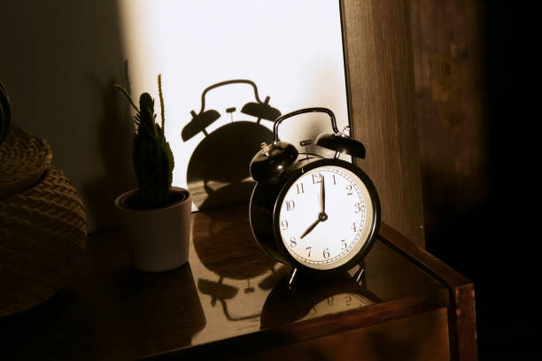 istock Black retro vintage alarm clock on a wooden bedside table with reflection in a polished surface in the rays of the setting sun with hard shadows 1148451990