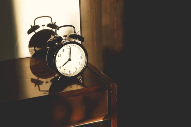 istock Black retro vintage alarm clock on a wooden bedside table with reflection in a polished surface in the rays of the setting sun with hard shadows 1148452008