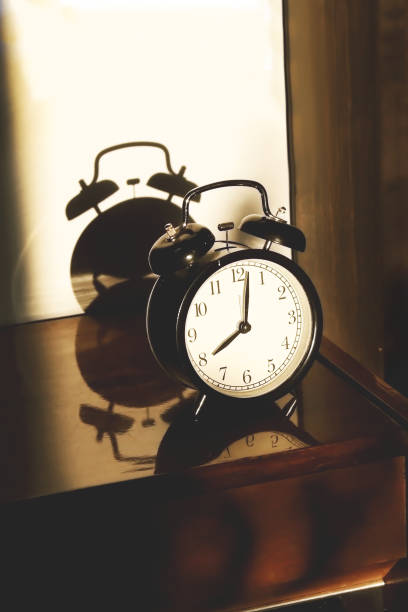 istock Black retro vintage alarm clock on a wooden bedside table with reflection in a polished surface in the rays of the setting sun with hard shadows 1148452027
