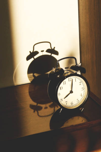 istock Black retro vintage alarm clock on a wooden bedside table with reflection in a polished surface in the rays of the setting sun with hard shadows 1148452031