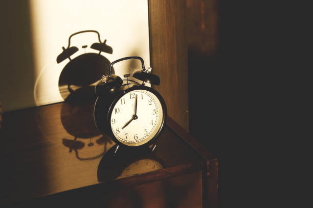 istock Black retro vintage alarm clock on a wooden bedside table with reflection in a polished surface in the rays of the setting sun with hard shadows 1148452033