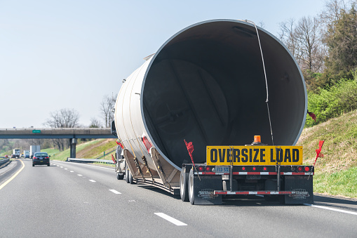 Woodstock, USA - April 18, 2018: Oversize load hauler truck trailer vehicle hauling concrete pipe tube on interstate highway road in Virginia with yellow warning signs and red flags