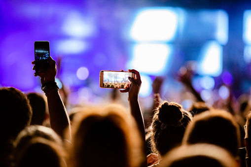 Unrecognizable fan taking picture of stage performers at music concert at night.