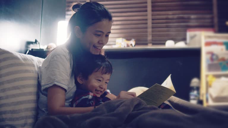 Asian Mother And Little Son Reading In Bed At Night.