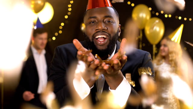 Glad african male blowing gold confetti dancing night club, holiday celebration