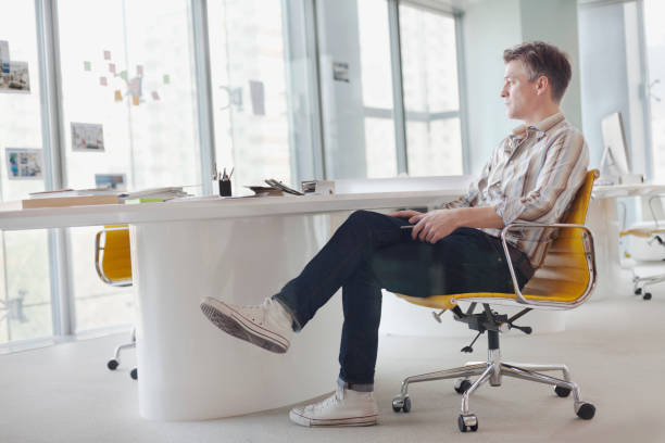 Mature adult creative type graphic designer interior architect product innovation think-tank studio owner entrepreneur startup small business sitting in chair looking out of window forward thinking of the future and planning decisions strategy choices thinking outside the box in contemporary modern daylight bright sunny studio co-working space in urban city downtown casually dressed alone solitude quiet moment gray hair dreams reminiscent dedication passion